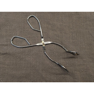May include: A silver-coloured metal kitchen tong with a scissor-like design, featuring two looped handles and curved gripping ends. The central pivot point is marked with the text 'Ekco' and 'USA' stamped into the metal surface of the tool.