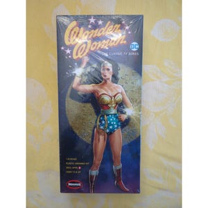 May include: A rectangular box for a 1/8 scale plastic assembly kit featuring Wonder Woman from the classic TV series. The packaging displays a graphic of the character in a red, gold, and blue costume against a dark blue background with stars.