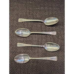 May include: Four silver-toned metal spoons arranged in a vertical column. Each spoon features an oval bowl and a flat handle with a decorative embossed border pattern along the edges. The metal surfaces exhibit a reflective, polished finish.