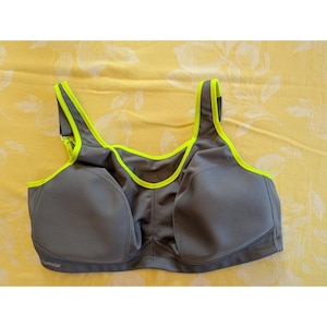 May include: A charcoal gray sports bra featuring bright neon yellow trim along the neckline and shoulder straps. The garment has molded cups, wide straps for support, and the brand name Glamorise printed in small letters on the lower left side.