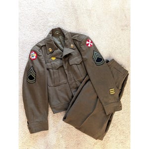 May include: An olive drab military uniform set consisting of a jacket and trousers. The jacket features two chest pockets, gold-tone buttons, and various embroidered patches on the shoulders and sleeves, including chevrons and circular insignia.