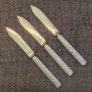 May include: Three silver-toned metal letter openers arranged diagonally. Each tool features a smooth, pointed blade and a handle decorated with an intricate, embossed floral pattern that covers the entire length of the grip area.