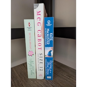 Puede incluir: Tres libros de bolsillo se muestran sobre una superficie de madera. Los libros se titulan "Sleigh Belles", "Size 12 and Ready to Rock" y "Daring in a Blue Dress". Las portadas de los libros presentan ilustraciones florales y retratos.