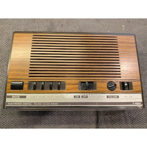 Puede incluir: Un radio reloj digital electrónico FM/AM rectangular con acabado de grano de madera. El panel frontal cuenta con una rejilla de altavoz horizontal, un botón de repetición grande, cuatro botones de control, un dial de volumen y un interruptor.
