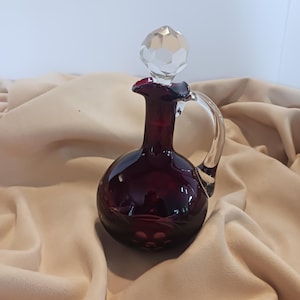 May include: A small, round, dark red glass decanter with a clear, faceted stopper. The decanter has a curved handle and a flared lip. The glass reflects light, creating highlights and shadows. The background is a neutral beige fabric.