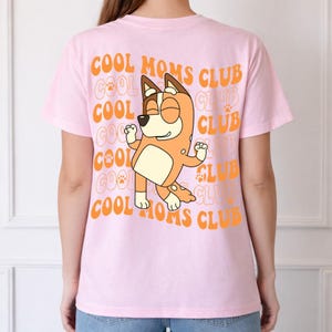 May include: A light pink t-shirt featuring a graphic on the back. The design includes an orange cartoon dog character with closed eyes and raised arms, surrounded by the repeating orange text COOL MOMS CLUB and small paw print icons.