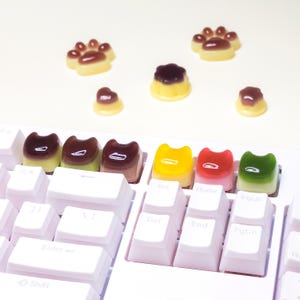 Può includere: Una tastiera bianca con tasti personalizzati a forma di gatto in vari colori, tra cui marrone, giallo, rosso e verde. Ulteriori accessori a forma di zampa di gatto e budino sono sparsi intorno alla tastiera.