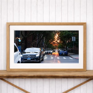 May include: A framed photograph of a city street at sunset. Cars are driving on the road, with a black Mercedes-Benz in the foreground. The sky is a mix of orange and yellow, and the trees line the street.