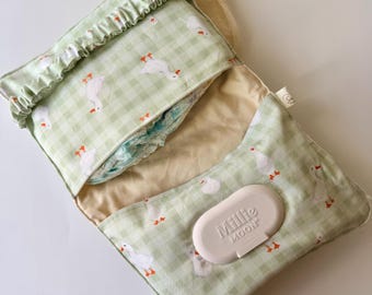 May include: A fabric diaper clutch featuring a light green and white gingham pattern with a repeating white goose print. The clutch has a ruffled top edge, a beige interior lining, and a beige plastic Millie Moon wipe dispenser.