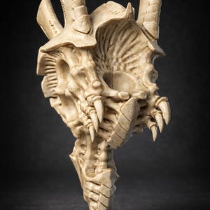 Peut inclure: Sculpture beige représentant une tête de créature avec des dents pointues, une mâchoire inférieure segmentée et des protubérances en forme de cornes. La sculpture texturée est placée sur un fond sombre.