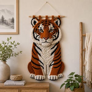 Op de afbeelding: Een gehaakte tijger wanddecoratie in oranje, zwart en wit. De tijger hangt aan een houten stok met een wit koord. De tijger zit rechtop. Een decoratief item voor in huis.