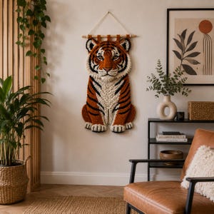 May include: A handcrafted tiger wall hanging. The tiger is made of yarn in shades of orange, black, and cream. The tiger is sitting upright with its paws forward. The wall hanging is suspended from a wooden dowel with a cream-colored string.