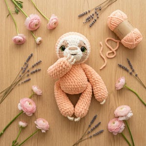 May include: A peach-colored crochet sloth toy with a white face and green eyes. The sloth is surrounded by pink flowers, lavender sprigs, and a ball of peach yarn on a wooden surface. The sloth is holding its hand up to its mouth.