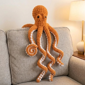 Może przedstawiać: Duża, pomarańczowa pluszowa ośmiornica z białymi akcentami na mackach. Ośmiornica ma czarne oczy z guzików i spoczywa na szarej sofie. Macki są długie i kręcone, ze spiralnym wzorem na jednym końcu.