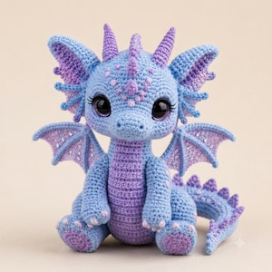 May include: A crocheted dragon toy featuring a light blue and purple color scheme. The creature has large black eyes with long eyelashes, pointed ears, small horns on its head, and wings with purple accents. A row of spikes runs down its back.