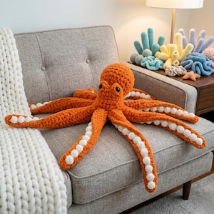 May include: An orange crochet octopus with white accents on its tentacles rests on a gray sofa. The octopus has black eyes and is surrounded by a white blanket. Decorative crochet coral and starfish are on a nearby table.