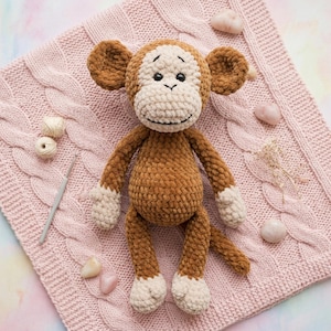 May include: A handmade brown and cream-colored crochet monkey doll with a smiling face, sitting on a pink cable-knit blanket. The monkey has white paws and a tail. Crochet hook and yarn balls are next to the monkey.