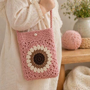 May include: A rectangular crochet crossbody bag in dusty pink featuring a central sunflower motif with a dark brown center and cream-colored petals. The bag has a single thin strap and a small white pearl-like button closure at the top center.