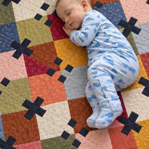May include: A patchwork quilt featuring a geometric pattern of squares in shades of rust, olive green, mustard yellow, pink, and blue, accented with dark navy cross-shaped fabric pieces throughout the design and visible quilted stitching.