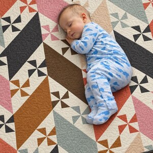 Peut inclure: Une courtepointe en patchwork présentant un motif géométrique de losanges et de moulins à vent dans des tons de rose, marron, gris et blanc, avec des coutures matelassées complexes sur toute la surface du tissu, offrant un aspect texturé.