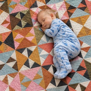 Op de afbeelding: Een patchwork quilt met een geometrisch patroon van driehoeken in tinten roze, mosterdgeel, terracotta, donkergrijs en lichtblauw. De quilt heeft zichtbare stiksels over het gehele oppervlak van de stof.