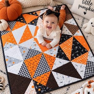 Könnte beinhalten: Eine rechteckige Babydecke mit einem geometrischen Patchwork-Design aus Dreiecken in Orange, Schwarz, Weiß und Grau. Die Stoffmuster zeigen Skelette, Spinnennetze, Fledermäuse und Karomuster. Die Decke ist mit einer schwarzen Einfassung versehen.