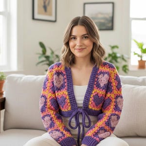 May include: A cropped, open-front crochet cardigan in a granny square pattern. The sweater features a purple border and tie, with squares of yellow, pink, and purple, each with a heart motif. The sleeves are long and the cardigan is worn by a person.