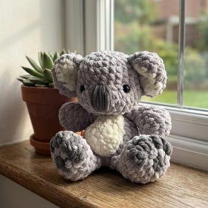 Puede incluir: Un peluche de koala de ganchillo gris y blanco hecho a mano. El koala tiene ojos negros, una nariz gris y detalles blancos en las orejas y el pecho. Está sentado en un alféizar de madera, con una planta suculenta en maceta en el fondo.