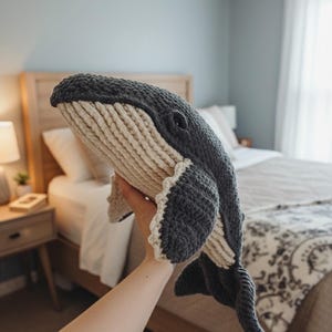 May include: A large, handmade crochet whale plushie in shades of gray and cream. The whale features a detailed design with a textured body, fins, and tail. The plush toy is held up in front of a bedroom setting.