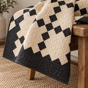 Könnte beinhalten: Eine Steppdecke mit einem geometrischen Schachbrettmuster in Schwarz und Creme. Der Stoff weist eine texturierte Steppnaht auf der gesamten Oberfläche auf, mit einem sich wiederholenden Quadratmotiv, das einen Kontrast erzeugt.