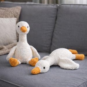 May include: Two white crocheted goose plush toys with orange beaks and orange feet. One goose is in a seated position, while the other goose is lying down in a horizontal position. Both plush toys feature small black eyes and textured yarn.