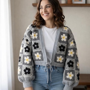 May include: A gray crochet cardigan with a floral pattern. The cardigan features square panels with white, black, and yellow flower designs. It has long, puffy sleeves and a tie closure at the waist. The person is wearing blue jeans and a white t-shirt.