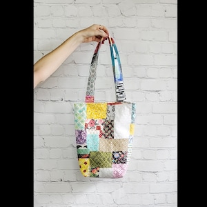 May include: A patchwork tote bag with colorful fabric squares in shades of yellow, green, pink, blue, and white. The bag has two long straps.