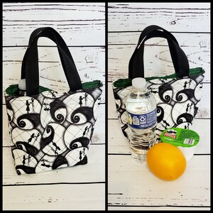 May include: A black and white lunch bag with a repeating pattern of Jack Skellington and Sally from The Nightmare Before Christmas. The bag has a black handle and a green lining. The bag is shown with a water bottle, an orange, and a container of Pringles.