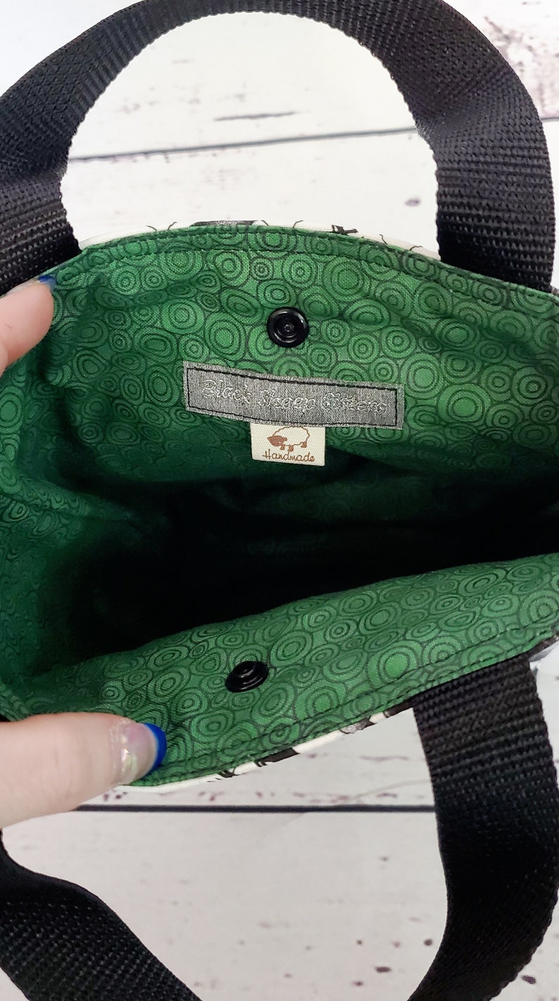May include: A green and white fabric tote bag with black straps. The inside of the bag is lined with a green fabric with a circle pattern. A label sewn into the inside of the bag reads "Black Bear Gifts" and "Handmade".