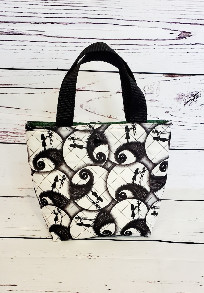 May include: A small black and white tote bag with a quilted design. The bag features a repeating pattern of silhouettes of Jack Skellington and Sally from the Nightmare Before Christmas movie. The bag has black handles.