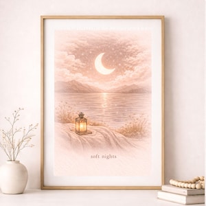 May include: A framed print depicts a serene night scene. A crescent moon glows above a calm lake, reflecting the moonlight. A lantern sits on a sandy shore, and the words "soft nights" are printed at the bottom. The color palette is soft, with peach and cream tones.