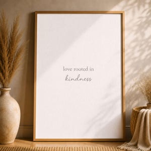 May include: A framed print with the words "love rooted in kindness" in a soft gray font. The frame is a warm gold color, and the print has a textured, off-white background. The image is styled with neutral decor.