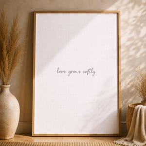 May include: A framed print with the text "love grows softly" in a delicate script. The frame is gold, and the background is a soft white. The print is displayed in a room with neutral tones and decorative elements.