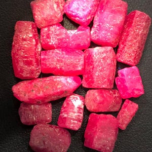 May include: A collection of raw, unpolished pinkish-red gemstone crystals with irregular, blocky shapes and textured, crystalline surfaces. The stones exhibit varying degrees of translucency and vibrant, saturated colour throughout their faceted structures.