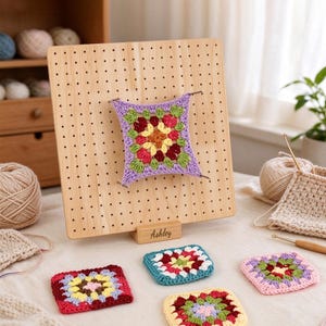 May include: A square wooden blocking board with a grid of holes and a base engraved with the name Ashley. A multi-coloured crochet granny square is pinned to the board with metal rods. Four additional crochet granny squares lie on the surface.