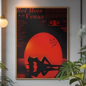 May include: A framed poster with the text "Red Moon in Venus" and a large red circle. The poster features a silhouette of a person reclining in front of the red circle. The artist's signature is visible. The poster is displayed on a wall.
