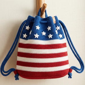 Peut inclure: Un sac à cordon de serrage crocheté à la main en rouge, blanc et bleu, ressemblant au drapeau américain. Le sac présente un haut bleu avec des étoiles blanches et des rayures rouges et blanches. Des cordons bleus servent de bretelles.