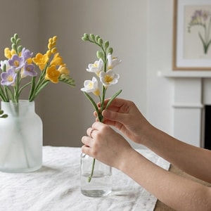 Op de afbeelding: Een persoon schikt een boeket van viltfreesia's in een heldere glazen vaas. De bloemen zijn in tinten paars, geel en wit. Andere vazen met soortgelijke bloemstukken staan op de tafel. Een botanische prent hangt aan de muur.