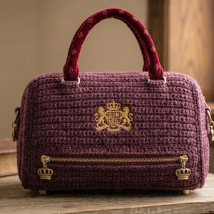 May include: A burgundy crochet handbag with a gold zipper and crown-shaped zipper pulls. The bag features a gold emblem and a burgundy velvet handle. The bag is sitting on a wooden surface.