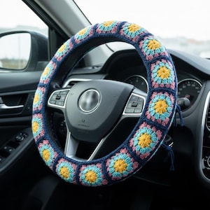 May include: A navy blue crochet steering wheel cover with a colorful floral pattern. The cover features square crochet blocks in shades of blue, yellow, and pink. The cover is shown installed on a car's steering wheel.