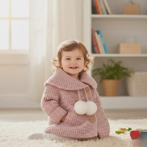 May include: A light pink crochet jacket with a hood and two white pom-poms. The jacket has wooden buttons and pockets. The child is sitting on a white rug. The jacket is a children's clothing item.