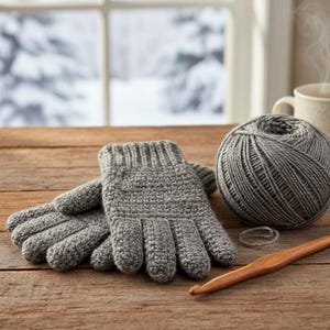 May include: A pair of gray, hand-knitted gloves with a matching ball of yarn, a wooden crochet hook, and a small ring. The gloves are on a wooden surface, with a window and snowy landscape in the background.