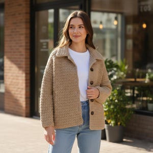 Könnte beinhalten: Beige Häkeljacke mit Kragen und Knopfverschluss. Die Jacke wird über einem weißen T-Shirt und mit blauen Jeans getragen. Die Textur der Jacke deutet auf ein handgefertigtes Produkt hin.