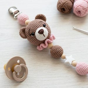 May include: A handmade crochet baby pacifier clip featuring a brown teddy bear head, pink details, and wooden beads. A beige dummy and crochet hook are also visible. The clip is designed to attach to a baby's clothing.